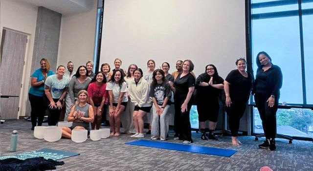 A large group of UTMB employees and students in yoga attire pose together; the instructor is seated in front of them with her singing bowls and a yoga mat nearby.
