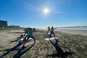Several yoga practitioners are standing warrior pose on mats on the beach, as the sun shines brightly