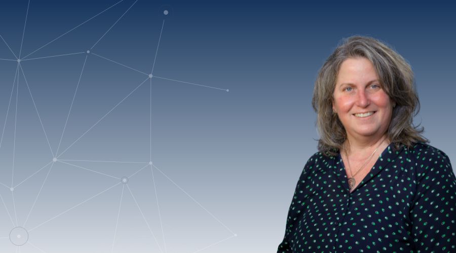 White female in her 50s with gray white wavy shoulder length hair , smiling, wearing a blue blouse with green dots with a pendant necklace.
