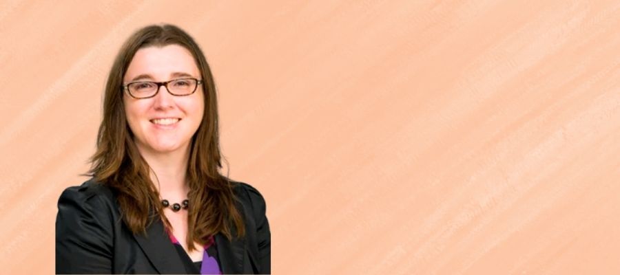 Elizabeth Lyons is a white woman in her 30s with long brown hair dark glasses who is smiling and wearing a black blazer and black beaded necklace.