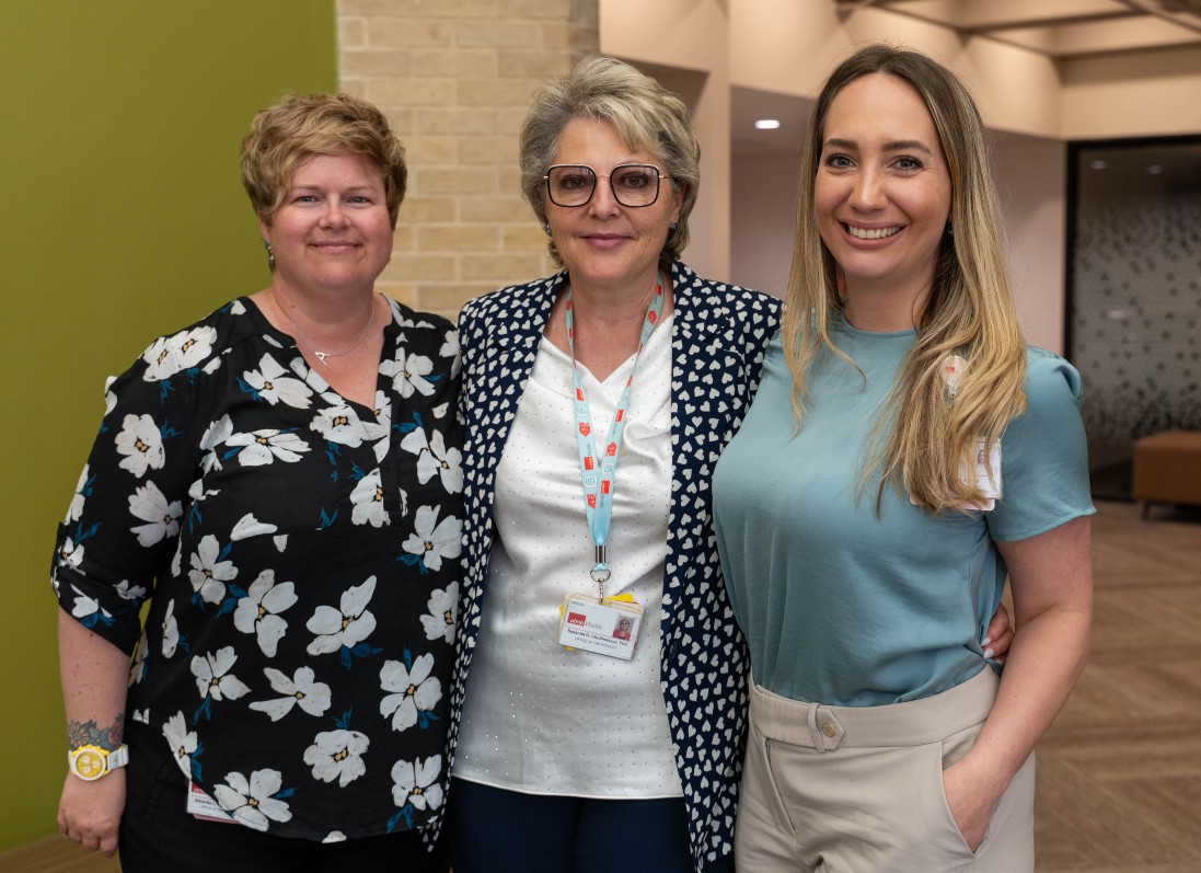 Three Caucasian woman standing closely. To the left, 40s, a bit heavier, has short  brown hair and in a floral blouse; next to her, 60s woman with light pixie hair and a white blouse. To the right, 30s woman, blond long hair, in a blue blouse.