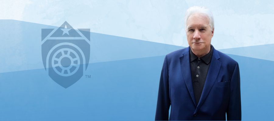 Presenter Karl Keller; White man in his early sixties with white hair and eyebrows with a fine line lip expression wearing a dark blue suit, buttoned, black collared shirt, standing half way looking forward.