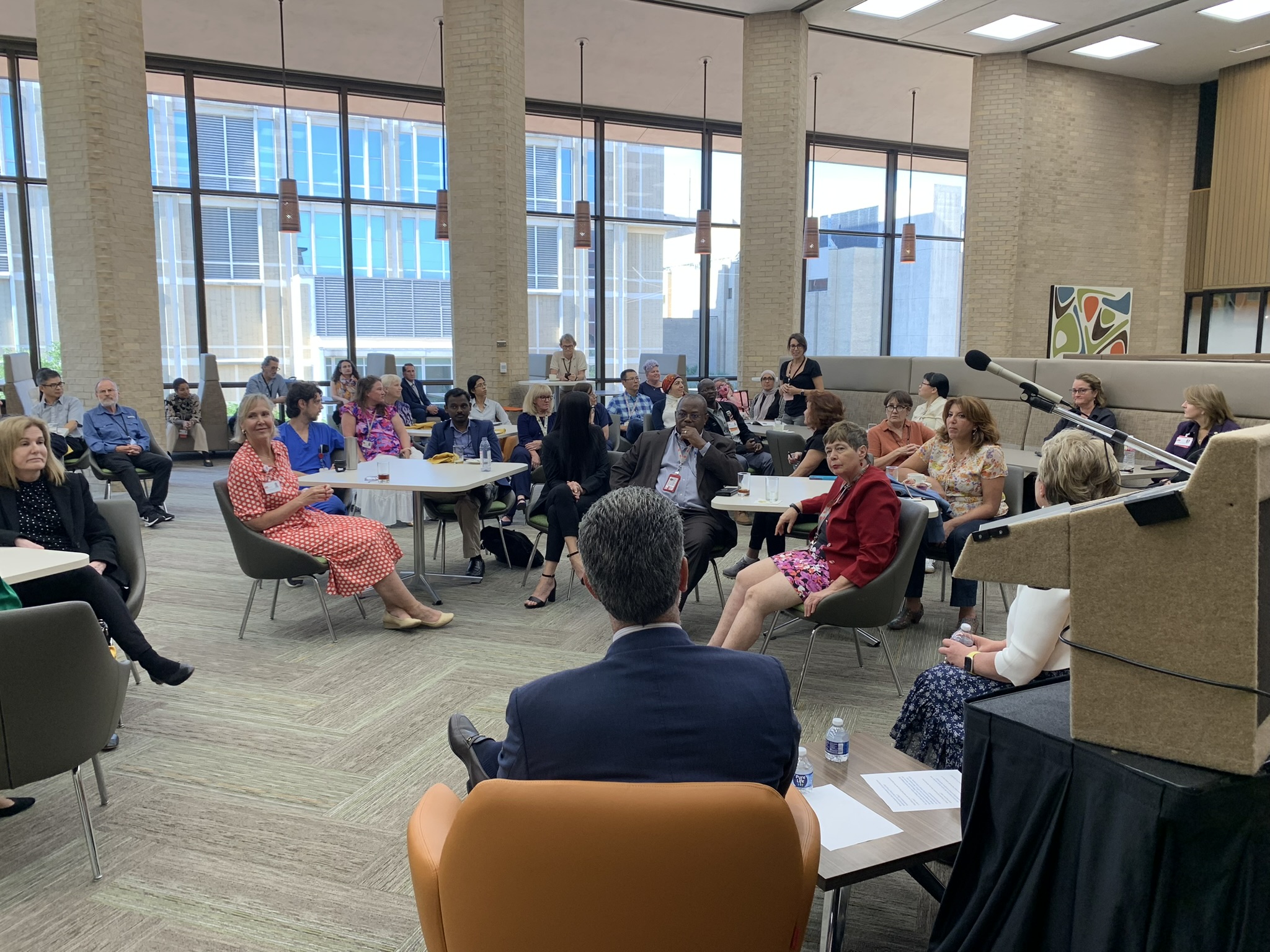 Open faculty lounge with windows from floor to ceiling against a wall, couch seating and tables and chairs with a variety of faculty members in formal attire seated.