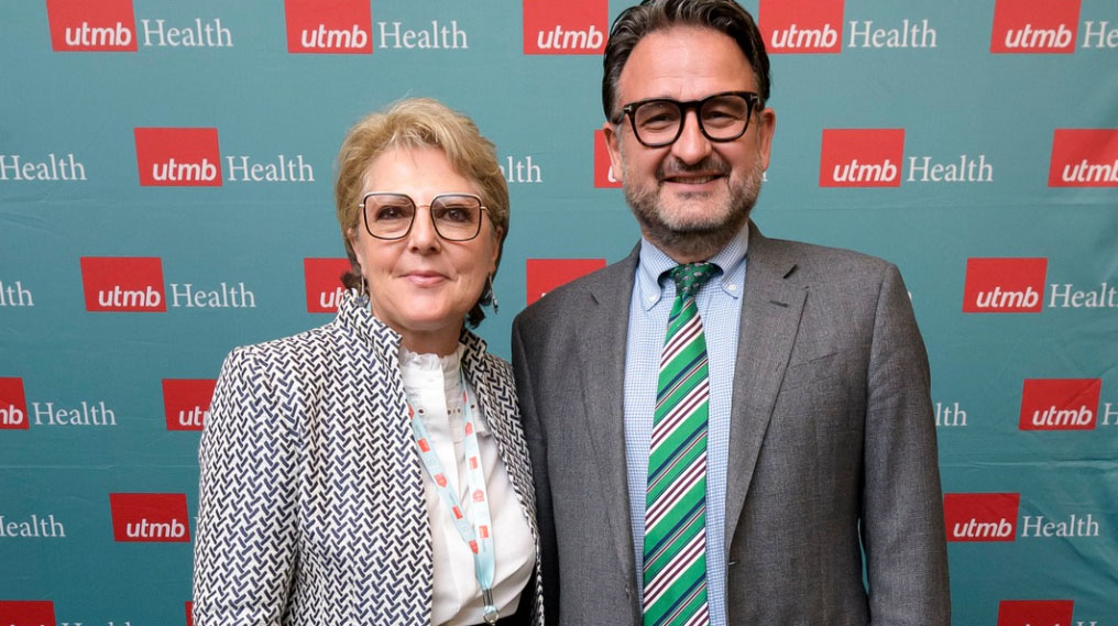 A Caucasian woman and man in front of a UTMB logo backdrop. The woman, in her 60s, with pixie light hair, black rimmed glasses and a patterned jacket. The man has short dark hair, dark rimmed glasses, in a gray suit with a green tie.