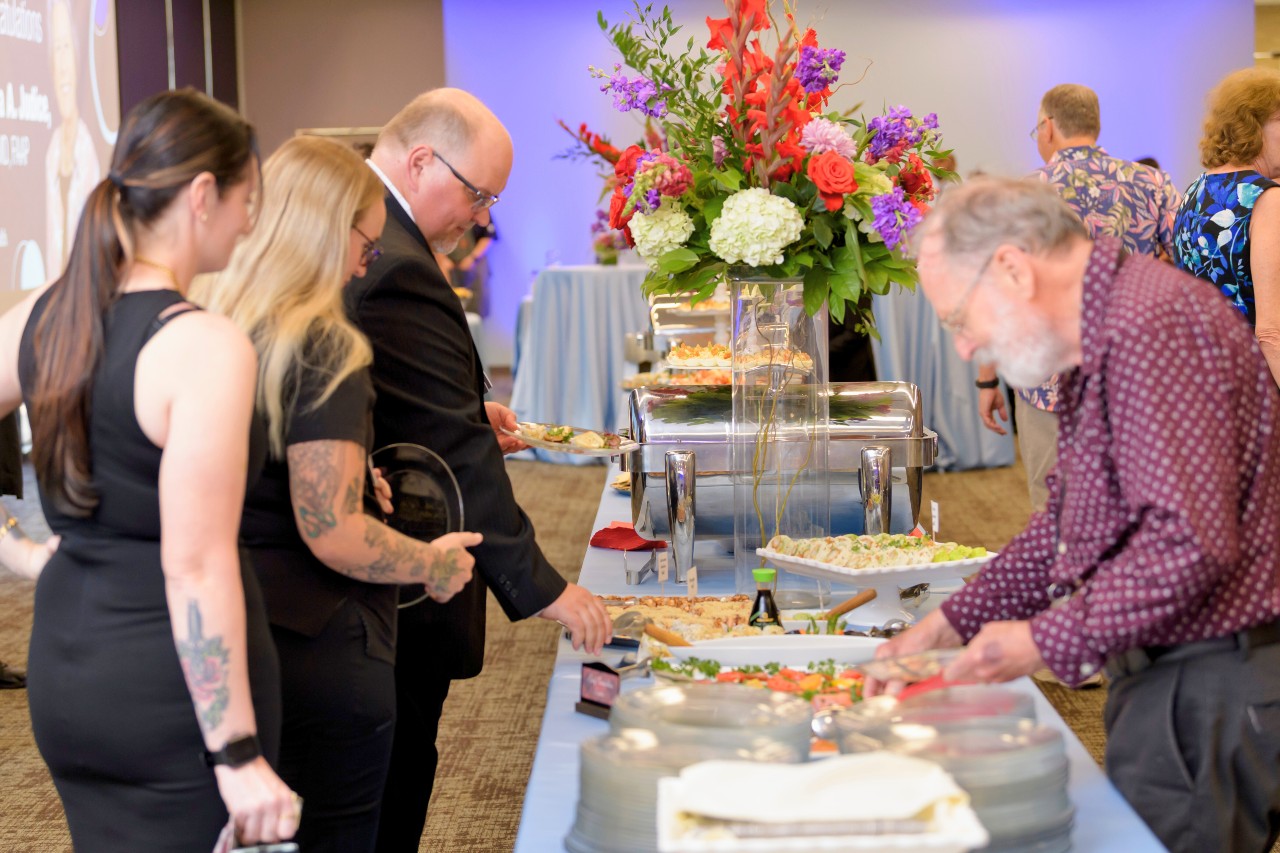 Event space with large buffet table with light blue table cover, multiple dishes with appetizers, a massive bright orange and purple floral arrangement and formally dressed attendees on both sides.