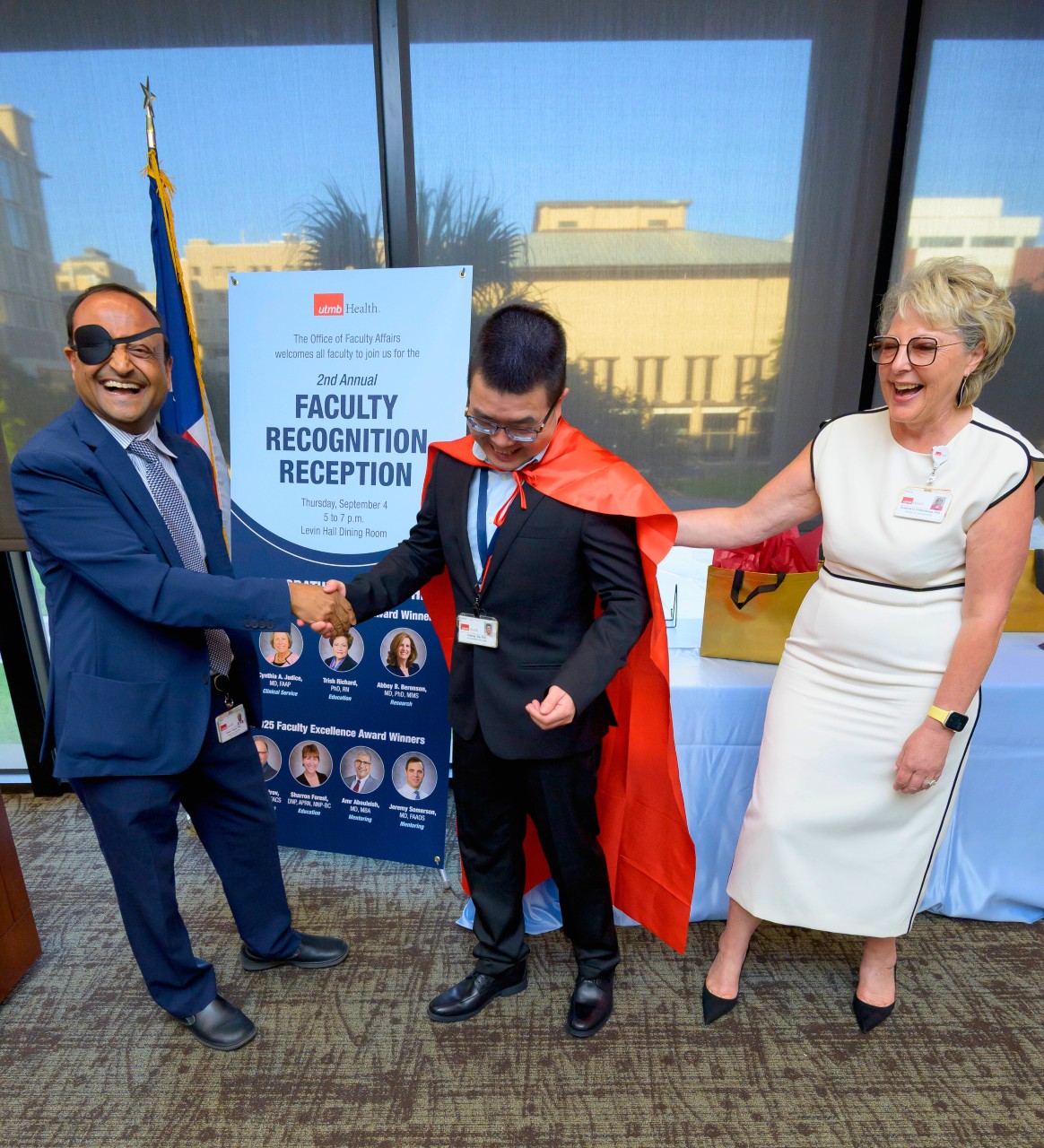An Indian man in his 50s in a vibrant blue suit with an eyepatch shaking hands with the Asian man in the center who is in a dark blue suit and cape. He is looking down.  A Caucasian woman in a white body fitted dress is patting his back. All are laughing.