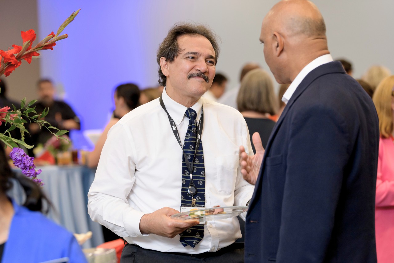 Two men of mixed ethnicities having a conversation; they are formally attired. One with his back facing with a dark blue jacket. The other with a white shirt and black tie with dark thinning hair and dark mustache.