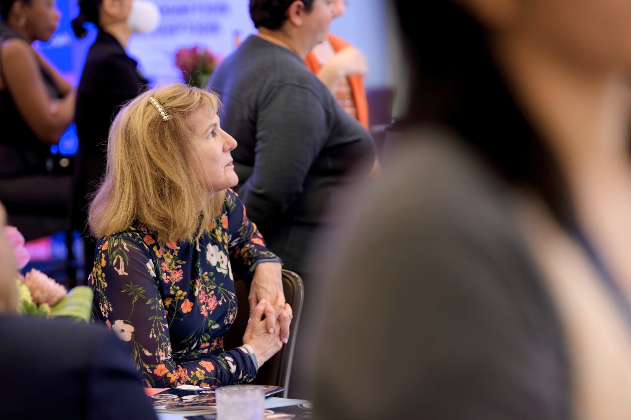 Profile of Caucasian female in her 50s with shoulder length blond hair looking at something, seated, wearing a dark blue long sleeved, floral dress with her hands clasped.
