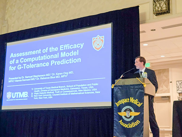 A speaker stands at a podium that has the Aerospace Medical Association name and logo on the front, as a screen to his right displays his presentation title, "Assessment of the Efficacy of a Computational Model for G-Tolerance Prediction"