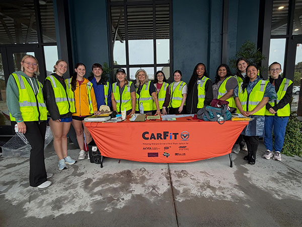 UTMB OT students wearing reflective yellow vests pose around a table with an orange tablecloth that says "CarFit"