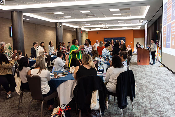 A busy room at the Clinical Partners Retreat, in which attendees mingle at tables and standing around the room, as someone speaks at a a podium