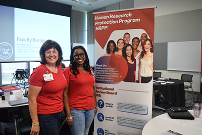 core showcase Two women in red shirts stand next to a banner that shares information about the Human Research Protection Program