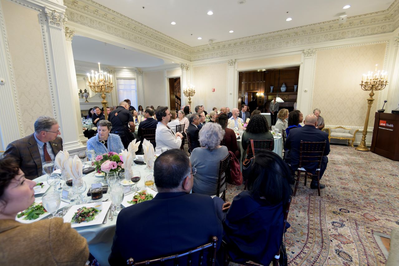 Large carpeted white walled event room with bordering trim with four visible round tables with six to eight  faculty seated at each wearing formal attire.