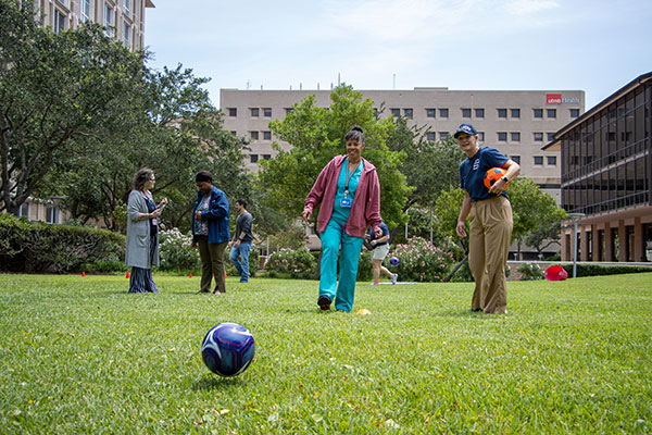 A woman in blue scrubs looks the soccer ball she just kicked on the lawn, as Wellness Director Julie Griffith watches, and other UTMB personnel stand nearby.