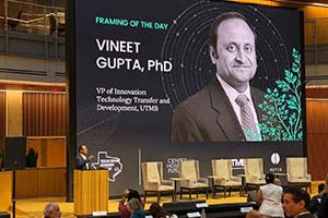 Dr. Vineet Gupta stands at a podium at the front of the summit as a large screen next to him shows his photo, name and topic title
