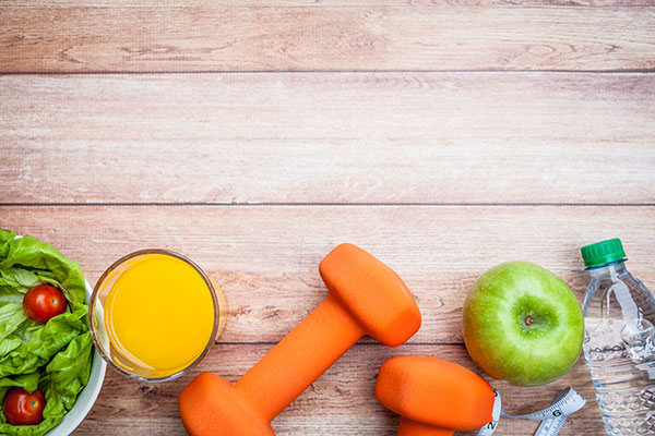 An aerial photo of a table with a salad, juice, dumbbells, an apple, tape measure, and water bottled lined along the bottom.