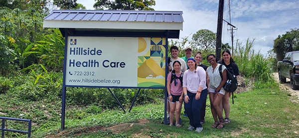 A group of nursing students and Dr. Zach Carson stand to the right of an outdoor sign for Hillside Health Care