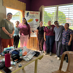 Nursing - Belize 4 A group of nursing students and staff from Hillside Health pose in a room in front of a health fair sign, with a table covered in supplies in the foreground.