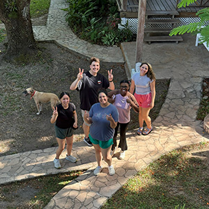 Nursing - Belize 7 A group of nursing students are outdoors looking upward toward the camera, some are giving the peace sign. There is a large brown dog next to them.