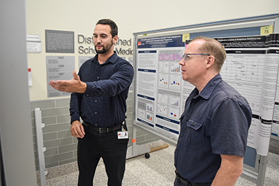 poster session 2 Two men are facing a mounted poster, one is holding his arm out to show the other something on the poster.