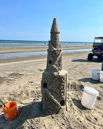 A tall, ornate sandcastle stands on the beach with a blue sky the background. Buckets and a golf cart surround the sandcastle.