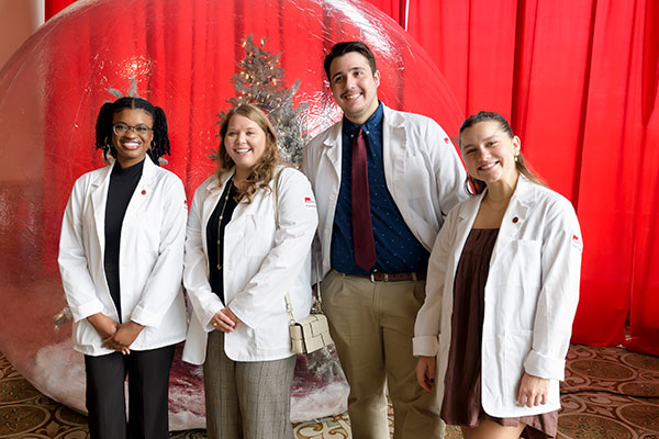 Four School of Nursing students in white coats pose in front of a red background with a Christmas tree at last year's Holiday Style event.