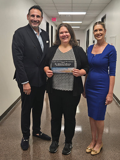 Tour de Blue Zones August Faculty Winner 2026 - sf Craig Kovacevich, left, and Tilly Clark, right, present the Blue Zones luncheon giveaway certificate to Dr. Rebecca Galloway, center.