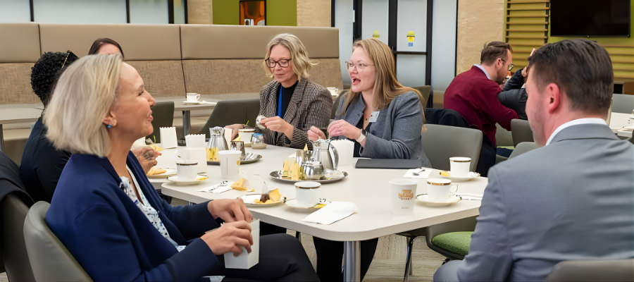 Faculty attending a tea networking session seated at a table. There are two blondish Caucasian women seated next to each other preparing tea cup with a tea set on the table and a Caucasian man and woman seated opposite them.