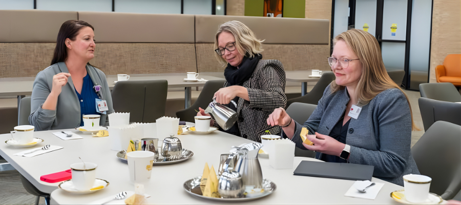 Three Caucasian women of 40-50s age seated at a table with tea service, filing cups and chatting. Two are blond and one is a brunette. All are dressed in business casual attire.