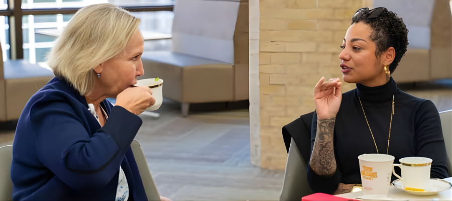 Two faculty members attending a tea event, seated at a table with cups and chatting. One is a Caucasian female with shoulder length blond hair in her 50s; the other is African American in her 30s with cropped short hair and gold hooped earrings.