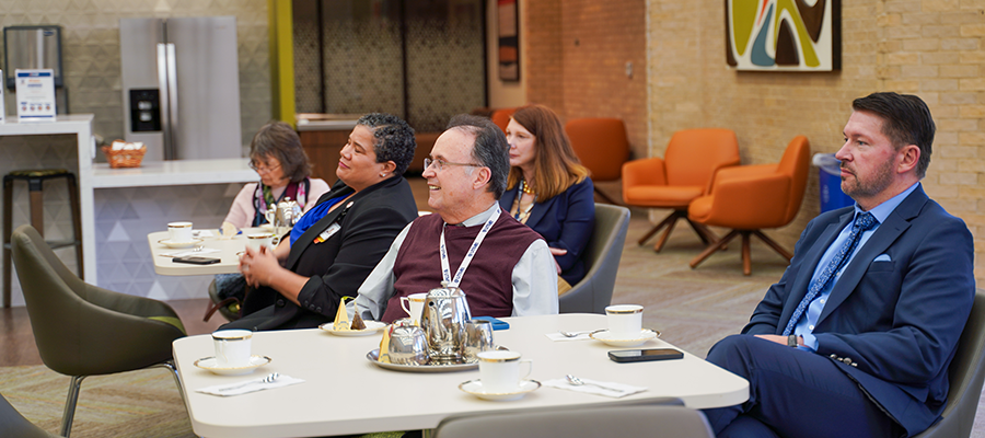 Faculty at two tables attending a tea and listening to a presented, seated profile. One man sits in the the back in a blue suit with a stoic expression. An older man at the same table wears a brown vest and smiles softly.