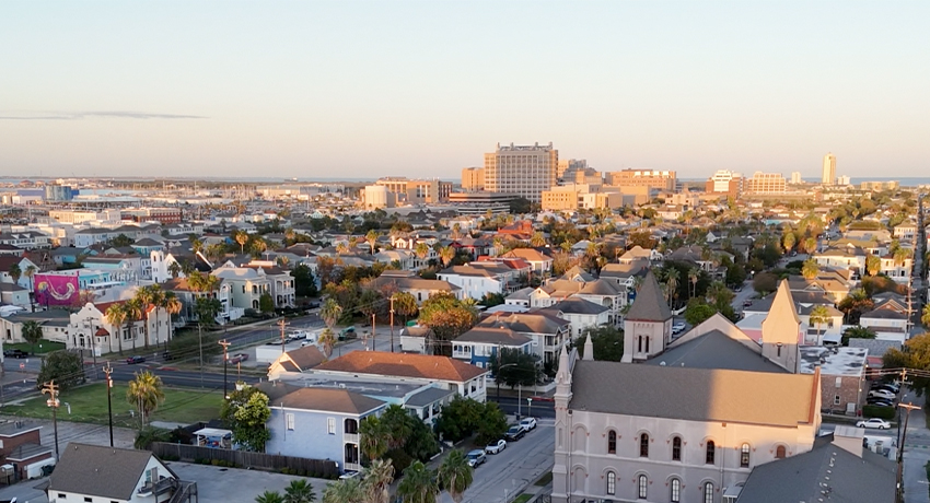 Galveston Island Featured Image A wide view of a coastal cityscape at sunset, showing rows of houses, tree‑lined streets, and several mid‑ and high‑rise buildings in the distance. The foreground includes a large building with towers casting long shadows.