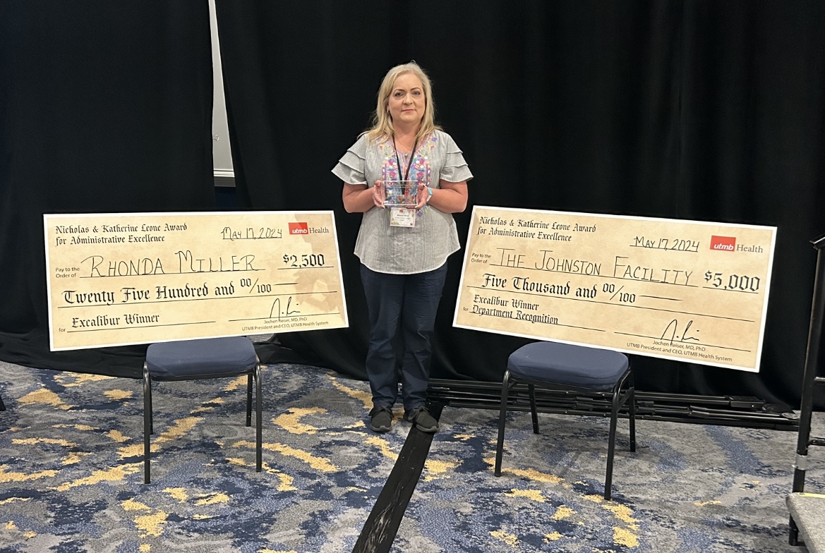 UTMB Employee Advisory Council Leone Award recipient standing on a stage holding a glass award, positioned between two large ceremonial checks labeled “Nicholas & Katherine Leone Award for Administrative Excellence,” with UTMB Health branding displayed.