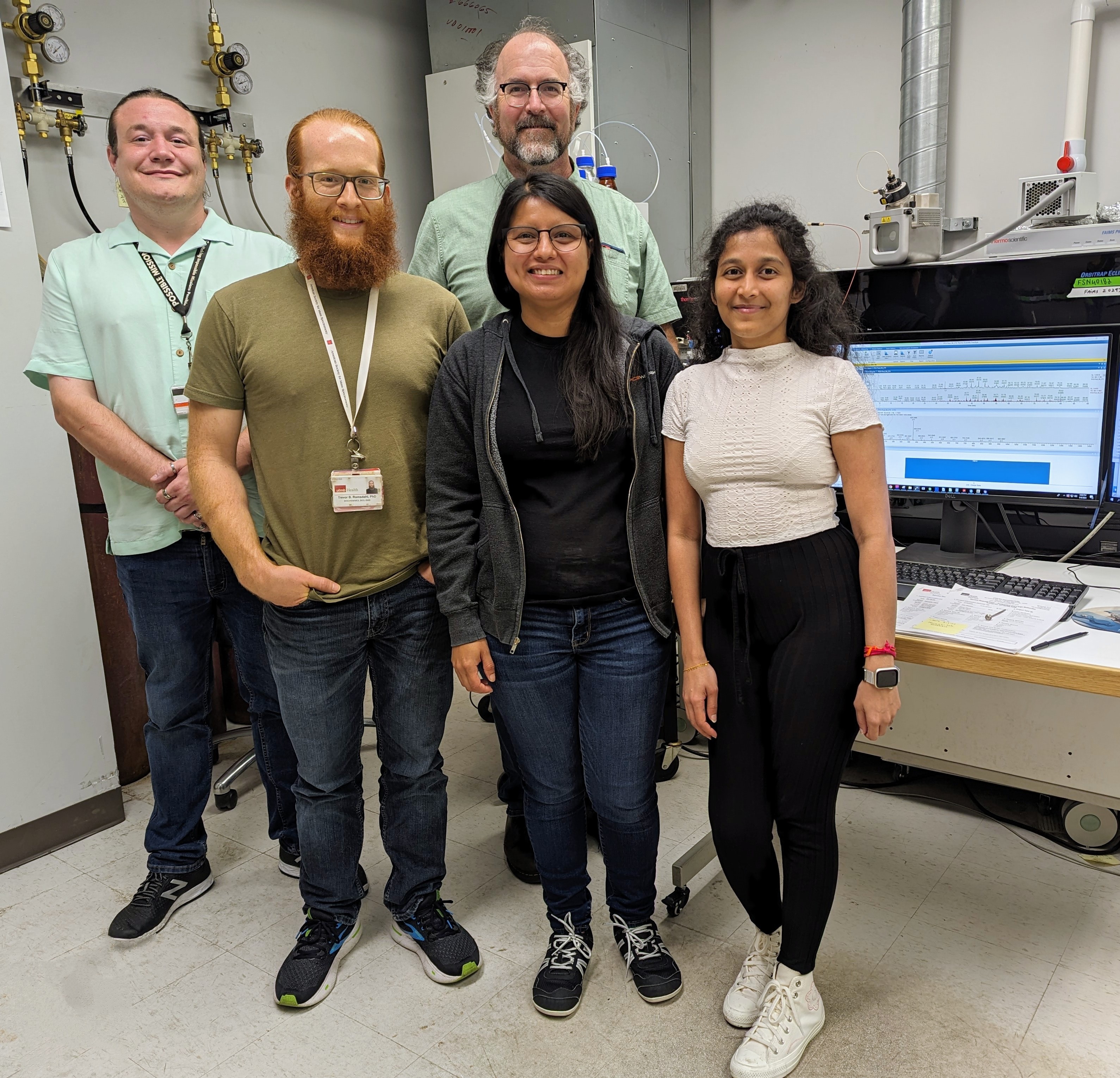 Five laboratory staff members standing together in a mass spectrometry lab beside analytical equipment and a computer displaying data.