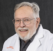 Picture of a man smiling wearing glasses and a dark colored shirt and white lab coat
