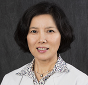 Picture of a woman smiling with short dark hair, wearing a white coat and brown and white spotted shirt
