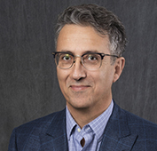 Picture of a man wearing glasses and smiling in a dark suit and purple shirt