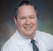 Picture of a man smiling wearing a white shirt, teal tie with white circles and a white lab coat