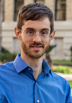 Picture of a man smiling wearing glasses and a blue shirt standing outside with a building in the back