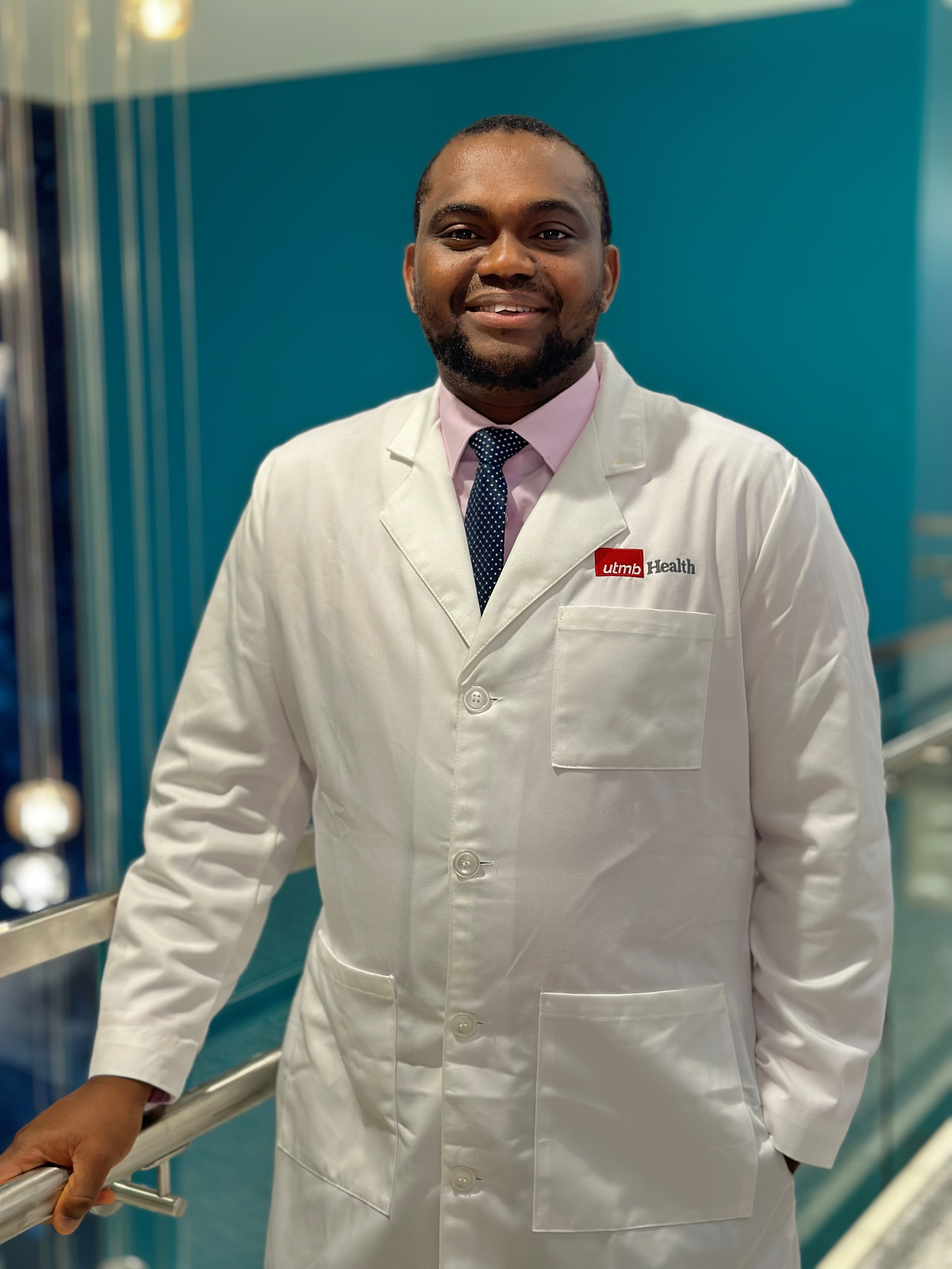 Portrait of a pharmacy preceptor standing indoors, wearing a white UTMB Health coat with a dress shirt and tie, against a teal interior background.