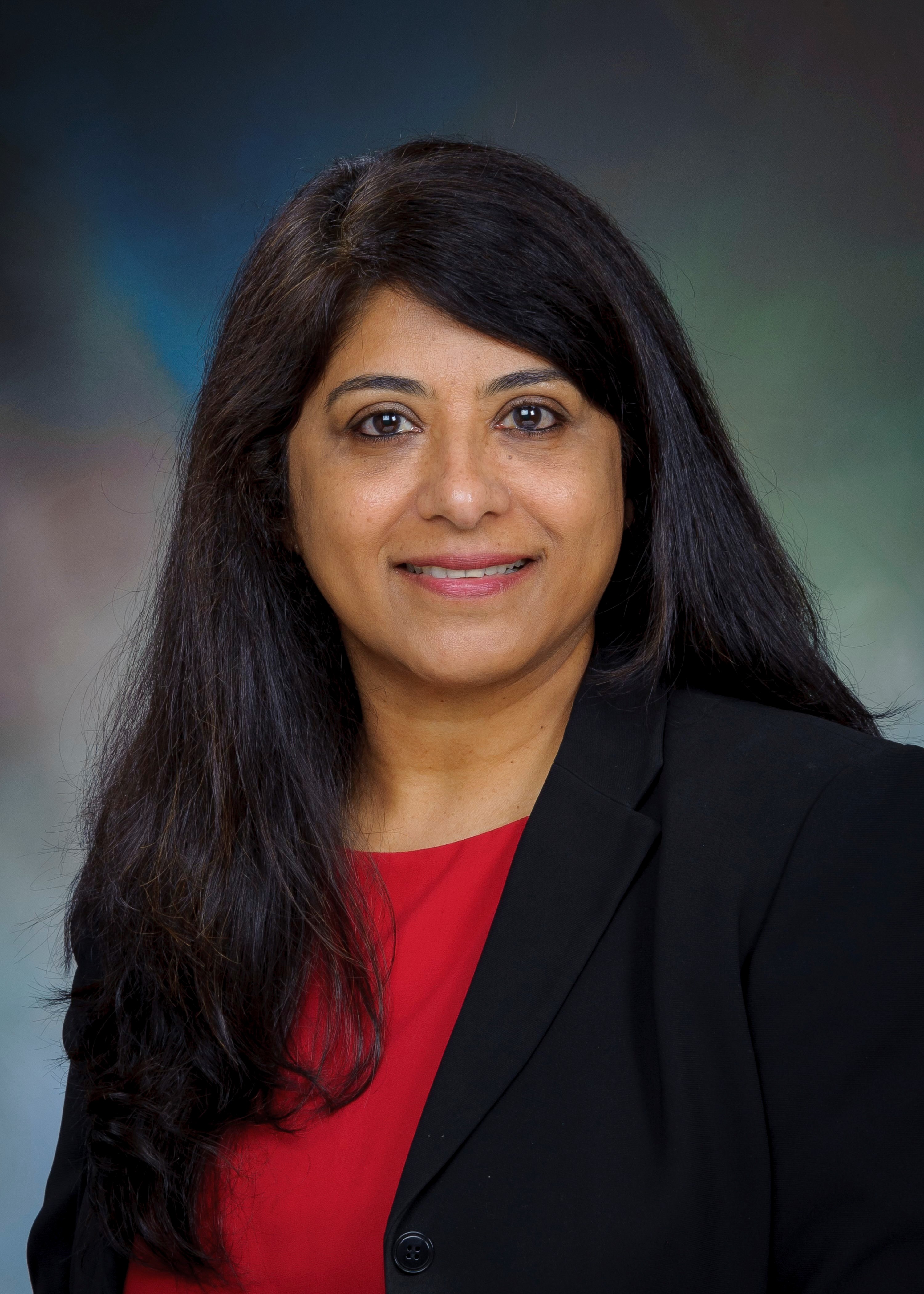 Studio portrait of a pharmacy preceptor wearing a black blazer over a red top, photographed against a softly blended neutral background.