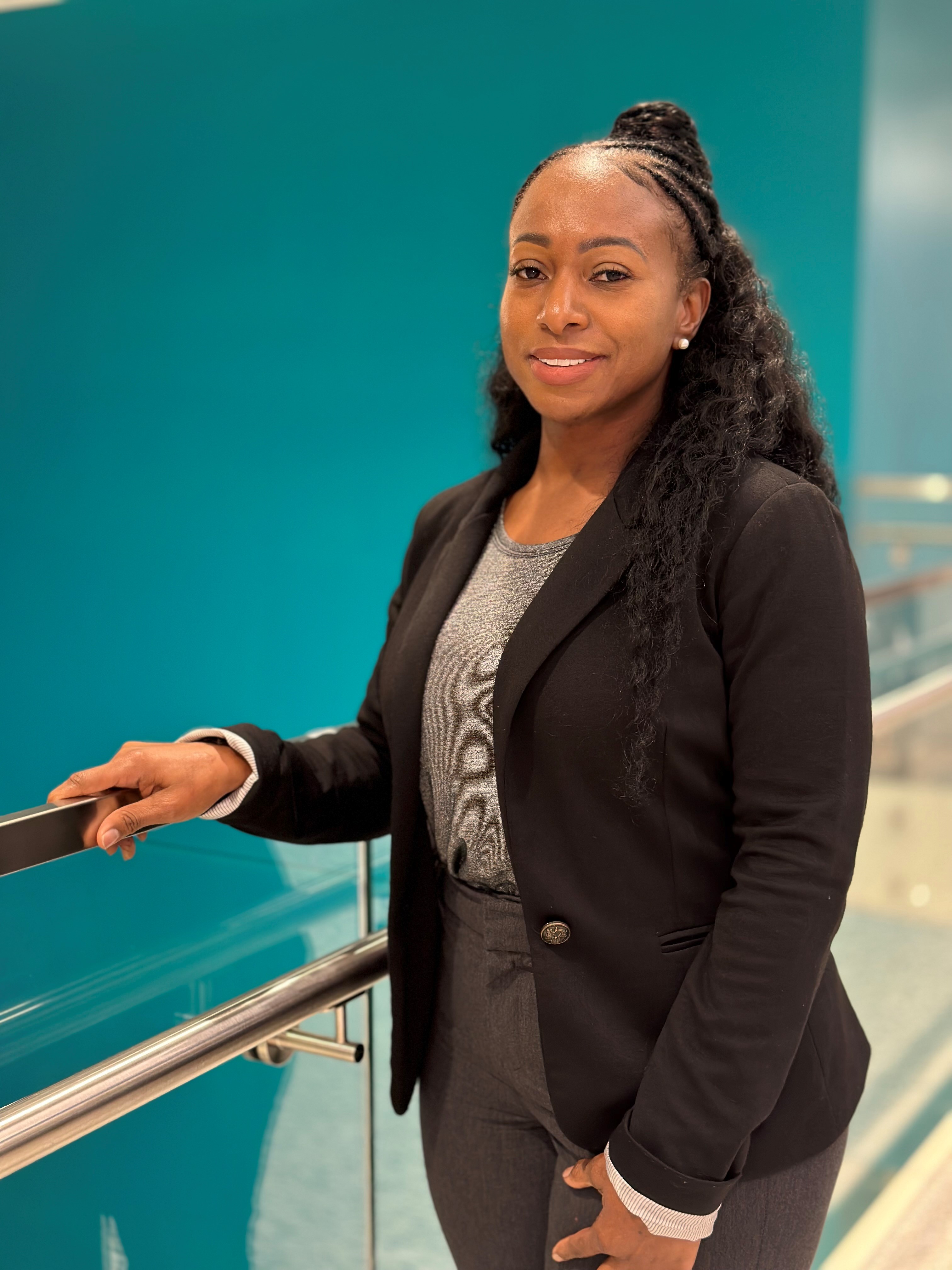 Portrait of a pharmacy preceptor standing indoors near a railing, wearing a black blazer over a gray top, with a teal interior background.