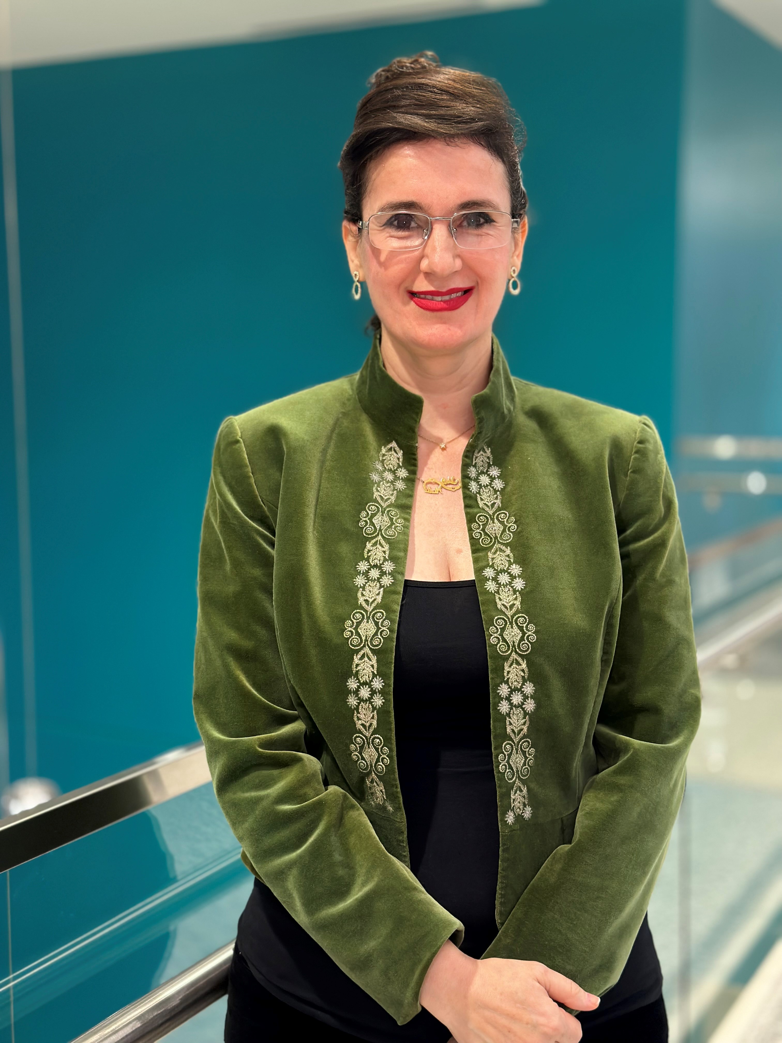 Studio-style portrait of a pharmacy preceptor wearing a green jacket with decorative embroidery over a dark top, standing indoors against a teal background.