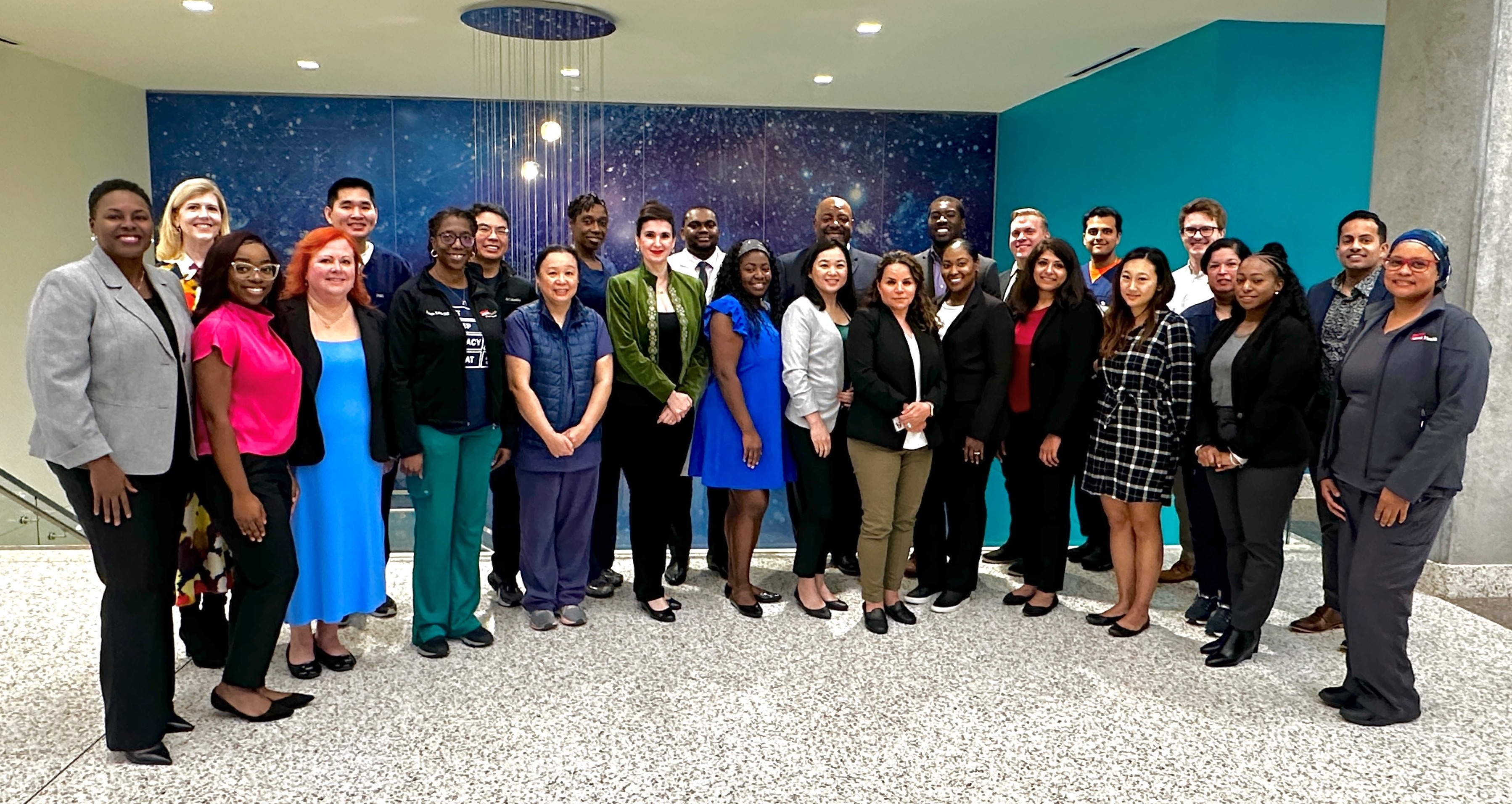 A large group of UTMB Pharmacy faculty, staff, and learners standing together indoors in a modern lobby with a blue and teal wall backdrop.
