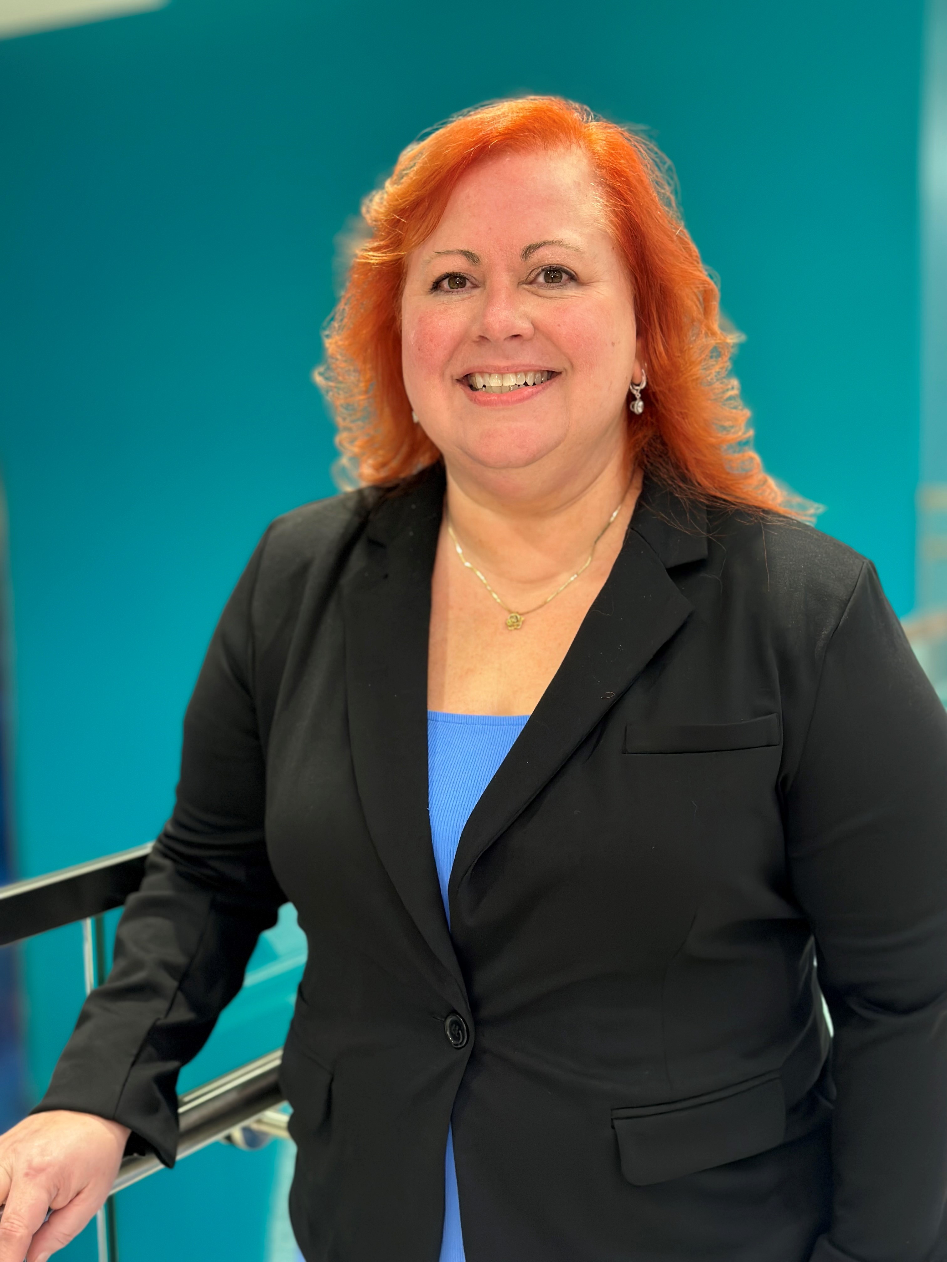 Portrait of a pharmacy preceptor standing indoors in a modern building, wearing a black blazer over a blue top, with a teal interior background.