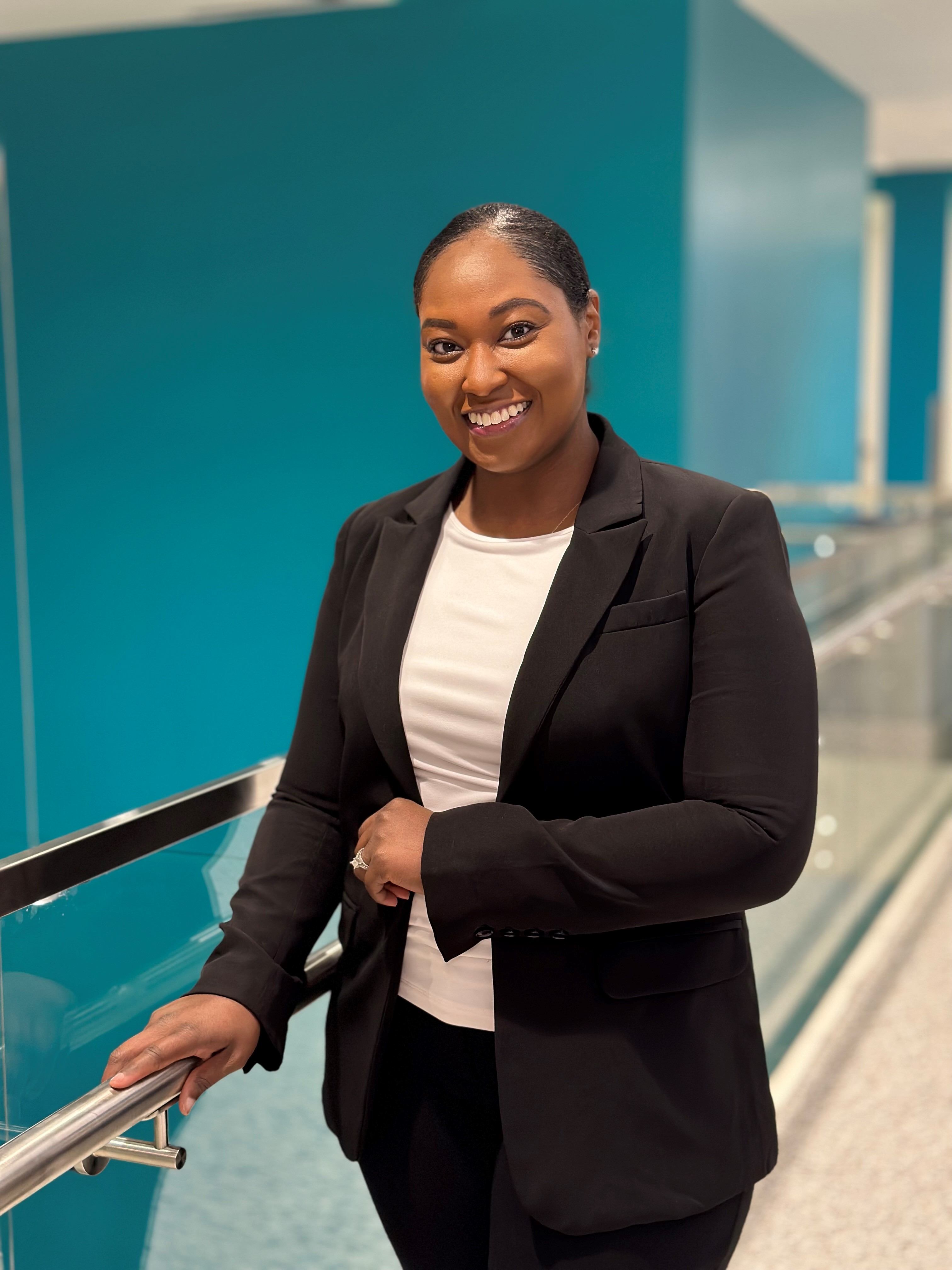 Portrait of a pharmacy preceptor standing indoors near a railing in a modern building, wearing a black blazer over a white top, with a teal interior background.