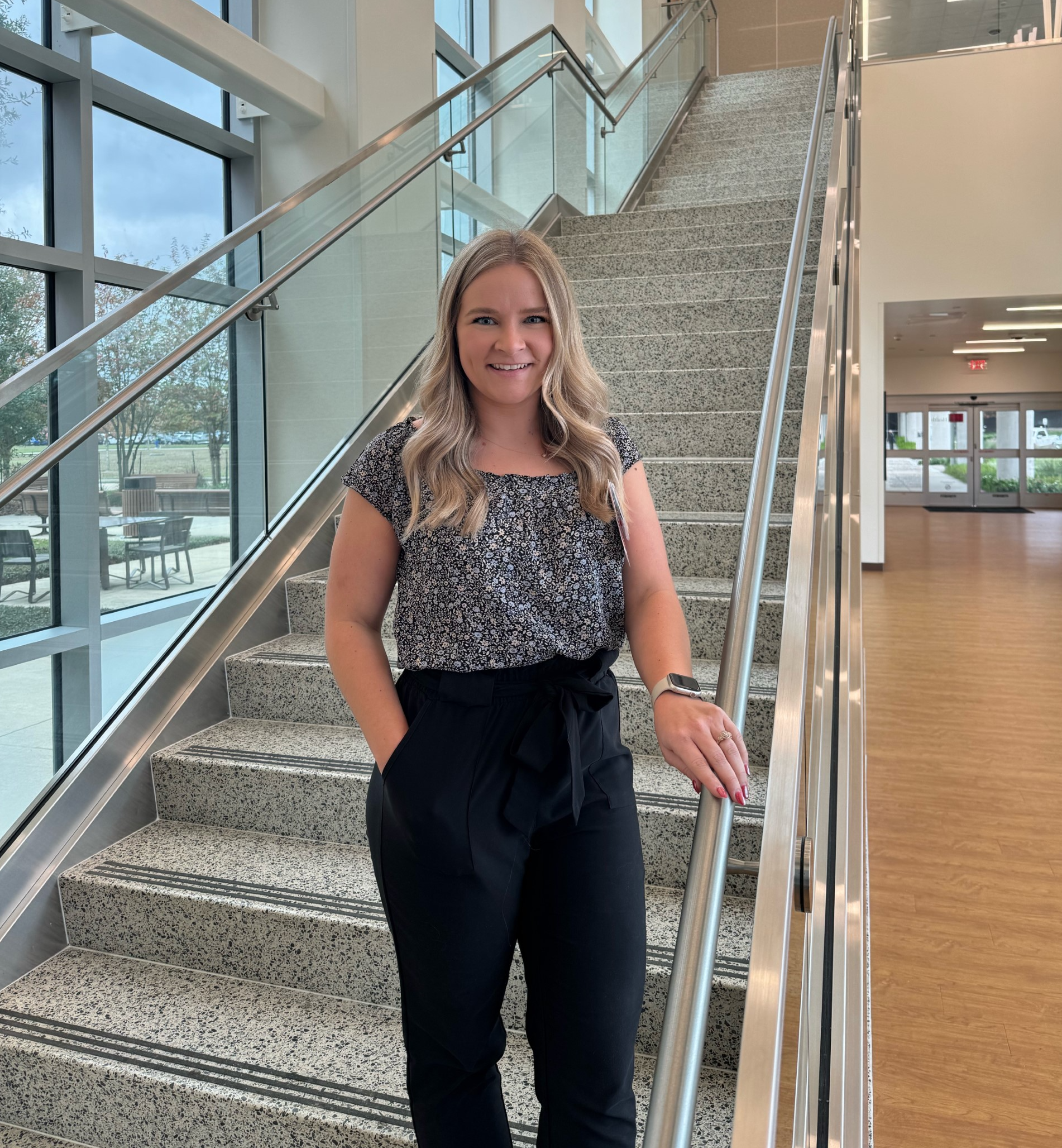 Portrait of a pharmacy preceptor standing on an indoor staircase in a modern building, wearing a patterned top and dark pants.