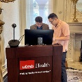 Two individuals at a UTMB Health podium adjusting a computer during a presentation setup in an ornate room with chandeliers, fireplace, and decorative molding.