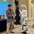 Two UTMB Alpha Omega Alpha attendees conversing in an ornate room with columns, candelabra lighting, and a display screen showing event planning information in the background.