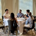 UTMB Alpha Omega Alpha attendees seated around a table sharing a meal and conversing in an ornate room with tall windows, columns, and patterned carpet.